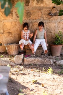 Two children sit on a stone bench against a rustic wall, surrounded by natural elements. They enjoy