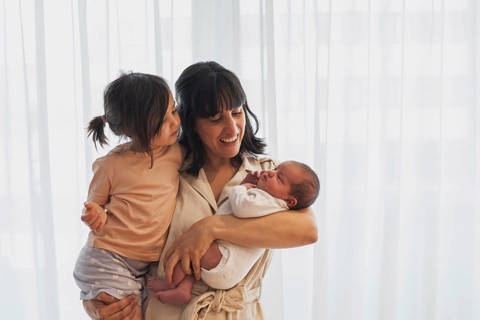 A joyful young family scene with a mother holding her newborn baby and big sister. Both children