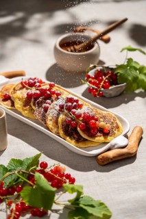 From above, a stack of golden pancakes is adorned with vibrant red currants and a sprinkle of