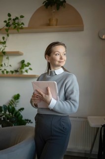 A woman stands in a cozy cafe, holding a tablet. She gazes thoughtfully towards the window. The