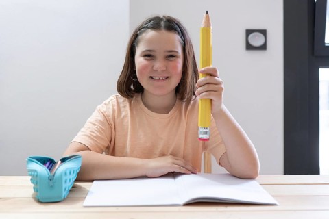 A smiling girl holds a giant pencil while sitting at a desk with an open notebook and a blue pencil