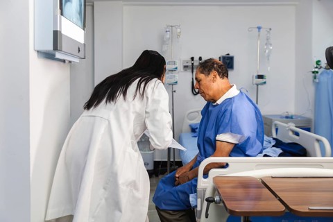 A female doctor in a white coat consults with an elderly male patient seated on a hospital bed,