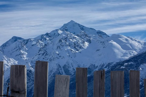 Majestic snowy peaks rise under a blue sky, framed by rustic wooden fence slats in the foreground,