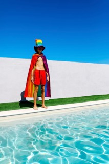 A person stands confidently by a pool under a blue sky, wearing a vibrant rainbow cape and hat. The