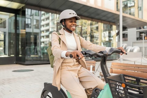 A joyful black woman rides a bicycle in a bustling urban area, wearing a stylish outfit and helmet