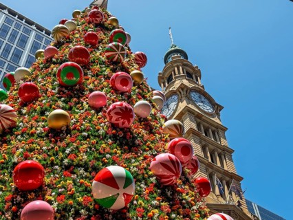 A beautifully decorated Christmas tree stands tall beside a historic tower under a clear blue sky