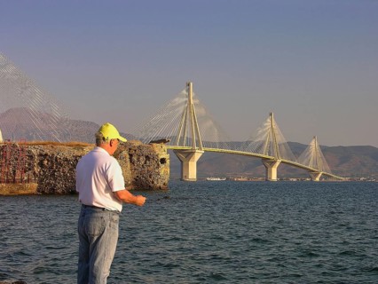 A man stands by the water and looks at a large suspension bridge, Rio-Antirrio cable car bridge,