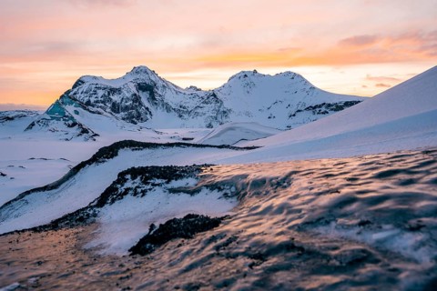 The snow-covered peaks of Katla Glacier glow under a soft orange-pink sunset, with textured