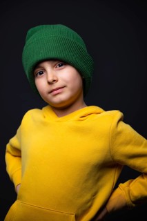 A child wearing a vibrant green beanie and a bright yellow hoodie confidently poses against a dark