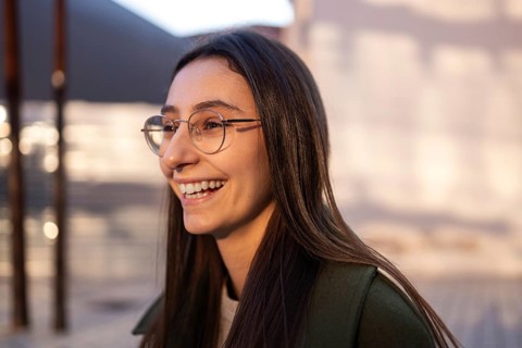 A woman with long hair and glasses smiles warmly, wearing a green coat Sunlight illuminates her