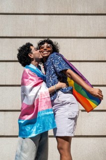 A multiethnic LGBTQ+ couple joyfully embracing, wrapped in vibrant Pride and transgender flags.
