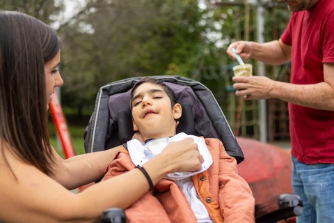 A family helps a her child with cerebral palsy in a wheelchair in an outdoor playground setting.