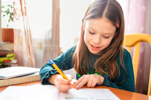 A young schoolgirl focuses on her homework at a wooden table, surrounded by books and illuminated