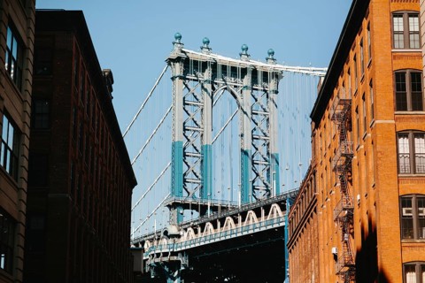 A striking view of a suspension bridge framed by two brick buildings in New York. The play of