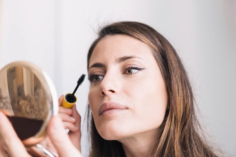 A woman applies mascara while looking into a mirror. Her face reflects concentration, highlighting