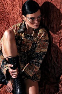 A stylish woman with slicked-back hair and tinted glasses poses fiercely against a textured