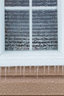 Close-up of glass pane window covered with thick sheet of ice and icicles hanging from edge of