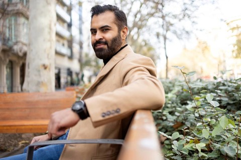 A cheerful Indian businessman in a stylish beige coat sits outdoors on a bench during a sunny