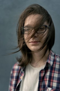A teenage boy with long hair and glasses stands against a gray background, wearing a casual plaid