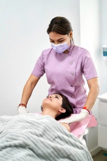 A beauty therapist in a lavender uniform and mask gently begins a facial treatment on a client,