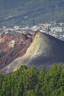 A volcanic hill of red rock rises in front of a town and dense pine forests, last volcanic eruption
