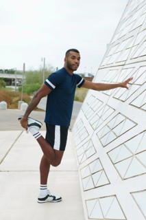 A black male athlete stretches his leg while leaning against a geometric wall. Dressed in sports