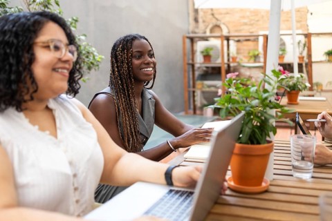 Multiracial women collaborate in an outdoor coworking space, engaging with laptops and notebooks.