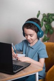 A young child wearing headphones studies at home using a laptop and notebook. The child is focused