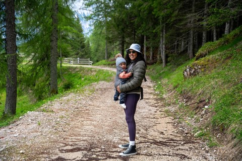 Mother carries her baby in her arms during a walk in a lush forest at the foot of the Dolomites.