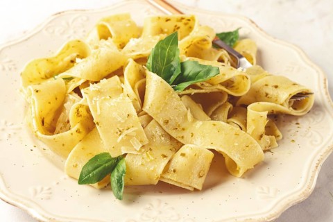 Fresh papardelle pasta, with basil and pesto sauce, is served, on a white plate, with a fork held