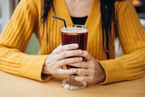 A woman wearing a yellow sweater holds a glass of a coffee with a straw at a wooden table The