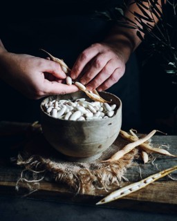 Close-up of hands shelling fresh white beans into a rustic wooden bowl, surrounded by empty pods on