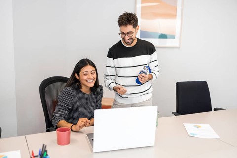 Two coworkers share a moment of laughter while collaborating at their office desk The image