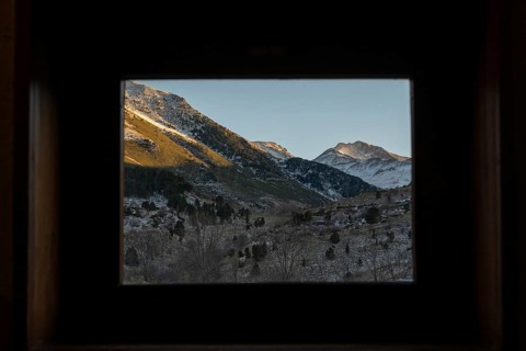A serene winter scene showcasing snow-capped mountains viewed through a dark, framed cabin window,