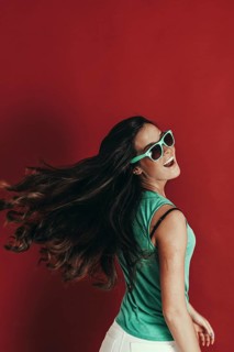 A woman with long hair, wearing green sunglasses and a teal top, smiles and turns her head