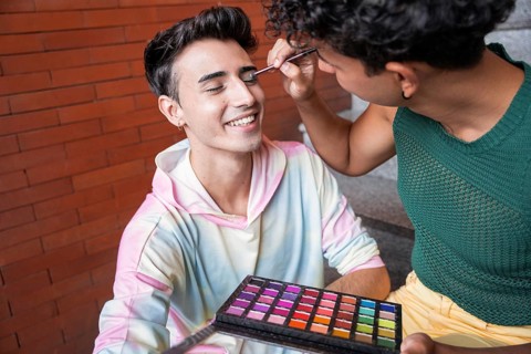 A happy couple enjoys a makeup session outdoors. One person applies colorful eyeshadow on the