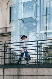 A woman in a stylish outfit walks along a modern urban pathway. The background features a glass