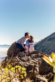 A couple shares a heartfelt kiss while sitting on top of a rocky mountain The sun is shining