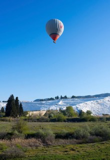 A hot air balloon floats over green countryside and white limestone terraces, sinter terraces, lime