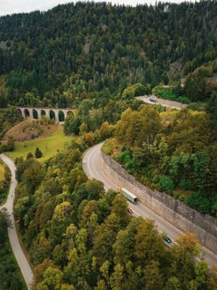 Aerial view of a winding mountain road lined with lush forest and a railway bridge in a hilly