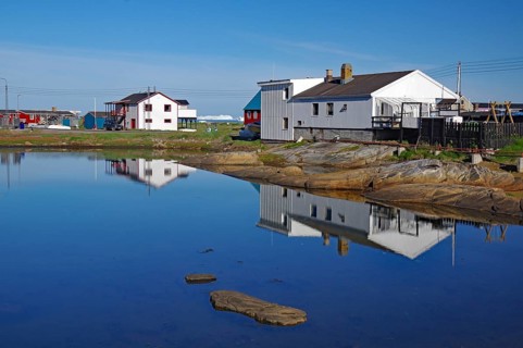 Simple houses are reflected in a calm lake surrounded by rocks and clear blue skies