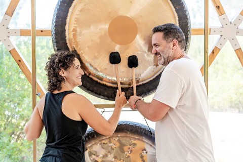 A man and woman, both sound therapists, are joyfully playing large gongs with mallets. They engage