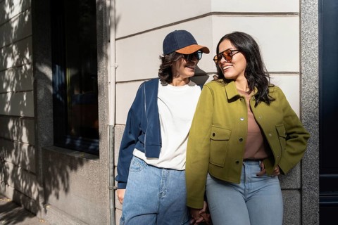 A happy lesbian couple enjoys a sunny day outside, leaning against a wall while holding hands. They