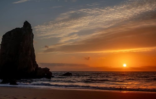 Scenic view of a towering rock formation at sunset, with the sun casting a warm glow over the ocean
