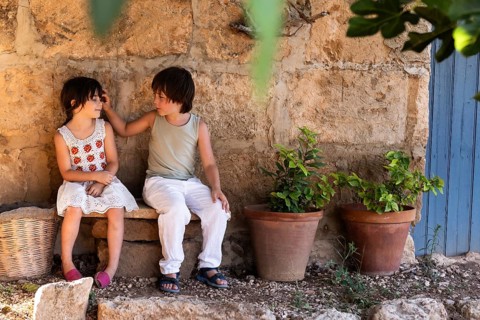 Two children sit on a stone bench by a rustic wall, engaging in conversation. They are surrounded