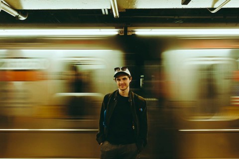 A man wearing a cap and jacket stands in a subway station with a blurred train moving in the