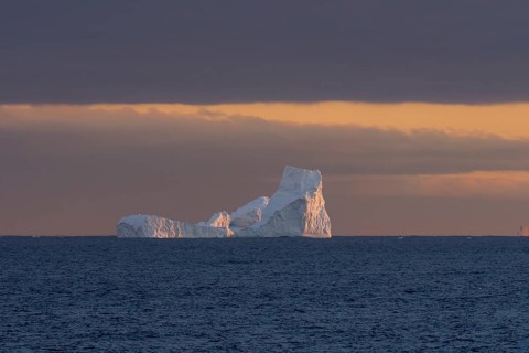 Iceberg in the open sea at sunset, warm light on ice structures, dark cloud cover on the horizon,