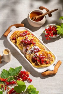 Top view of delicious pancakes topped with red currants on a white ceramic tray. The light linen