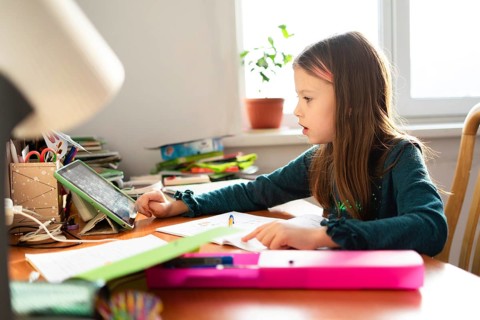 A young schoolgirl sits at her desk, focused on her homework. The scene includes a tablet and