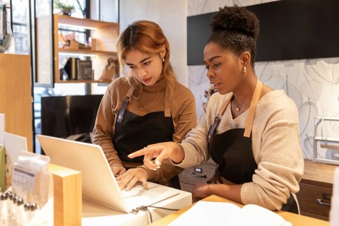 Two employees in a cosmetics store collaborate, discussing tasks at a laptop. They wear aprons,